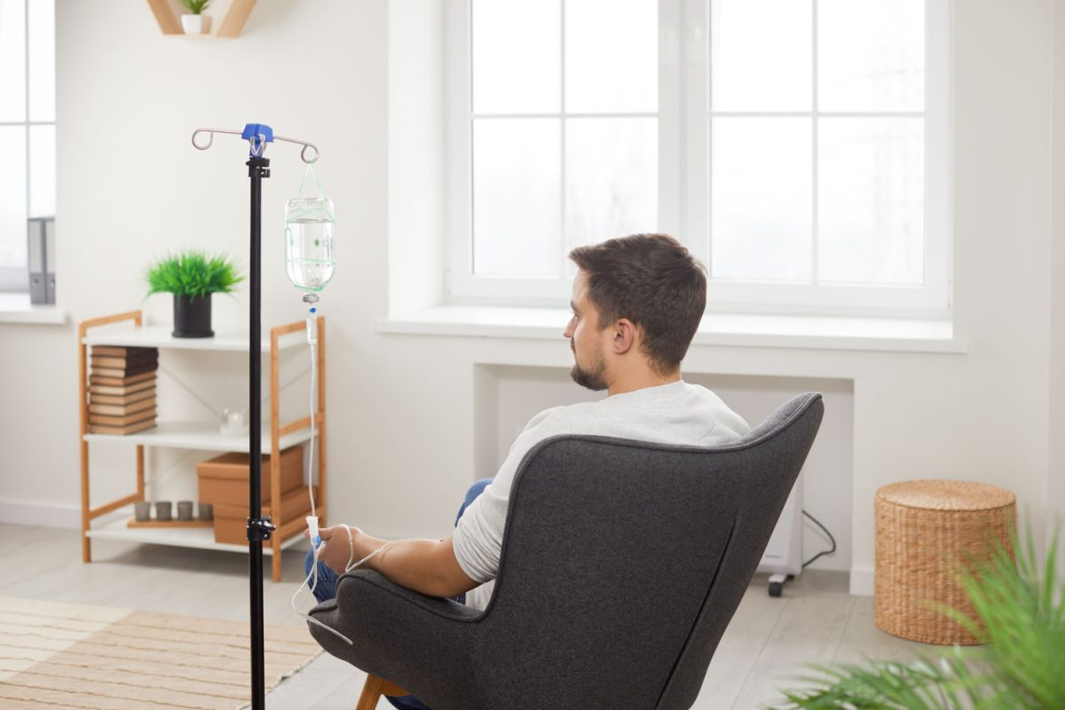 Rare view of male patient receiving intravenous treatment. Young man sitting in armchair during intravenous vitamin therapy in hospital room or wellness center. Medical treatment, healthcare concept