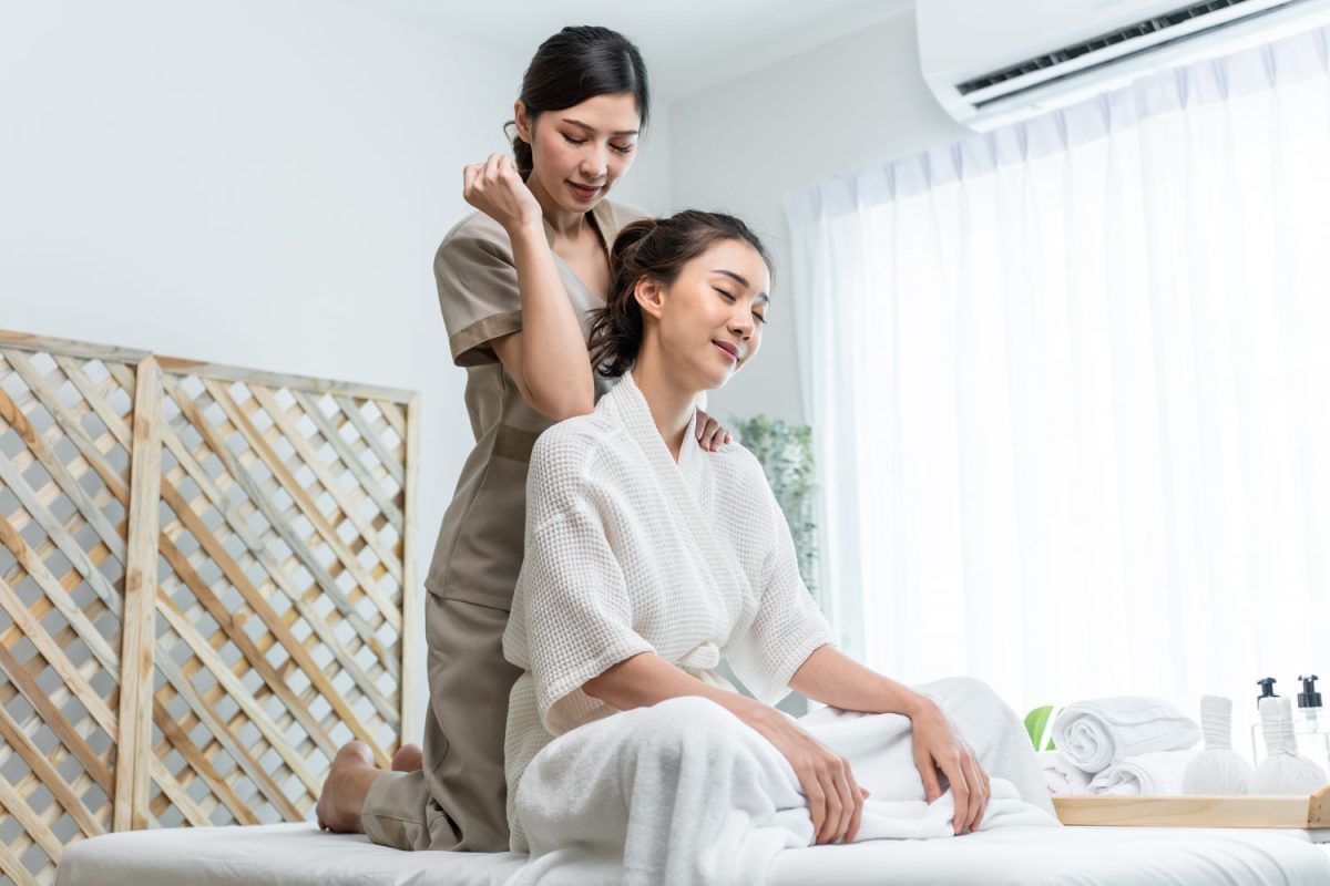 Asian young happy woman feeling relax during shoulder and back massage. Attractive beautiful girl sitting on massage table getting physiotherapy service from masseuse for body care in spa beauty salon